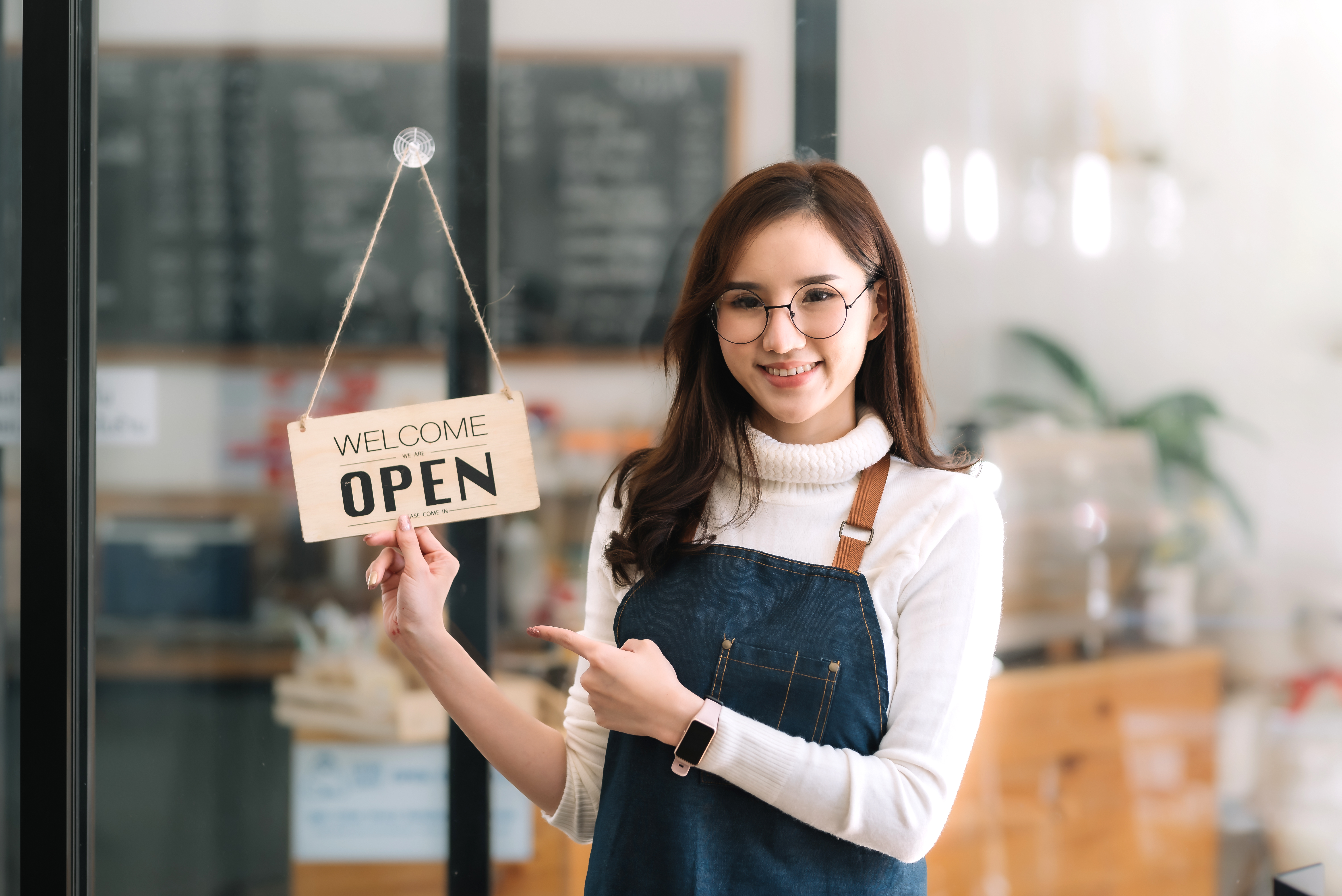 Happy young business owner woman standing pointing open sign board looking at camera.